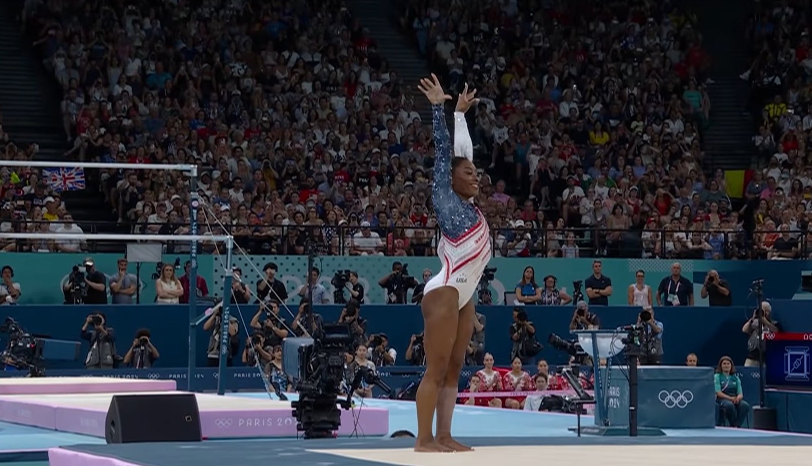 Simone Biles performs on a gymnastics beam, raising her hands in a poised position, showcasing balance and skill.