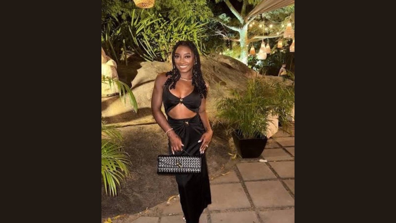 Simone Biles wearing a black dress and heels stands confidently, showcasing her elegant style and poise.