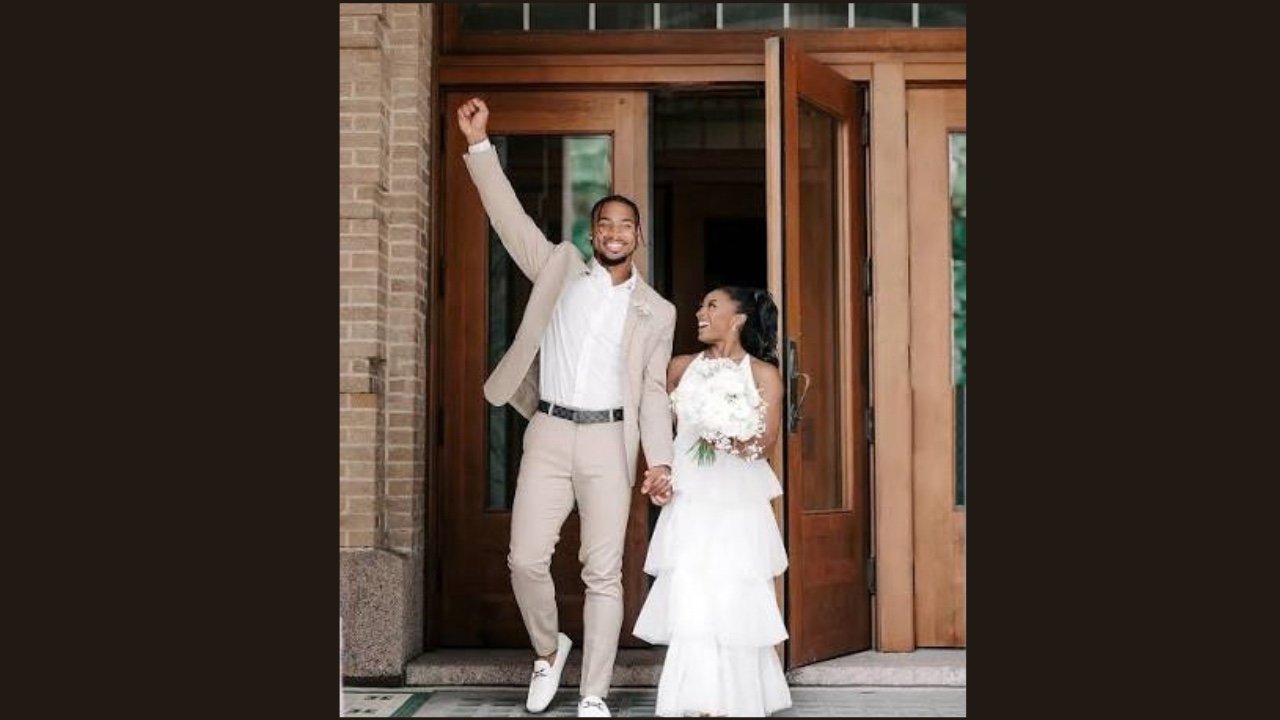 A bride and groom stand together outside a church, smiling and holding hands, surrounded by beautiful architecture.