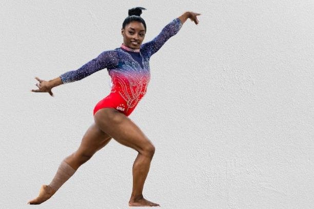 Simone Biles in a red and blue leotard performs a split on a gym mat, showcasing her flexibility and strength.