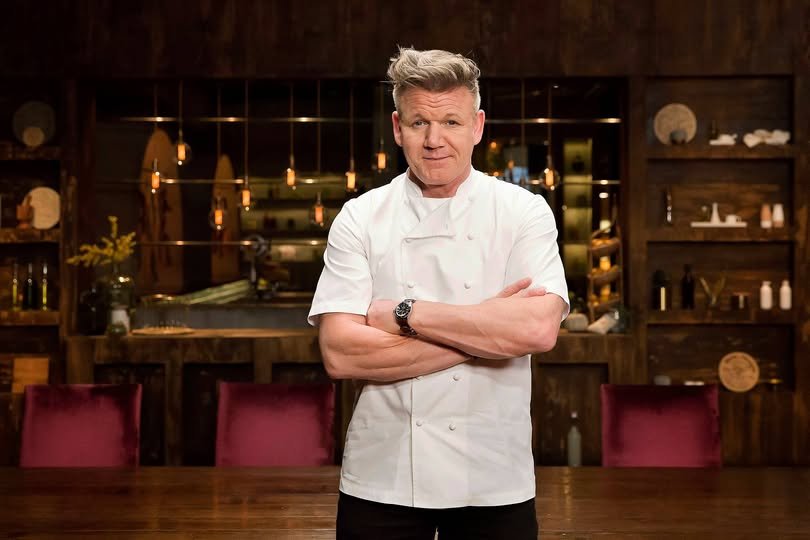 Gordon Ramsay stands confidently in front of a table, showcasing his culinary expertise and engaging presence.