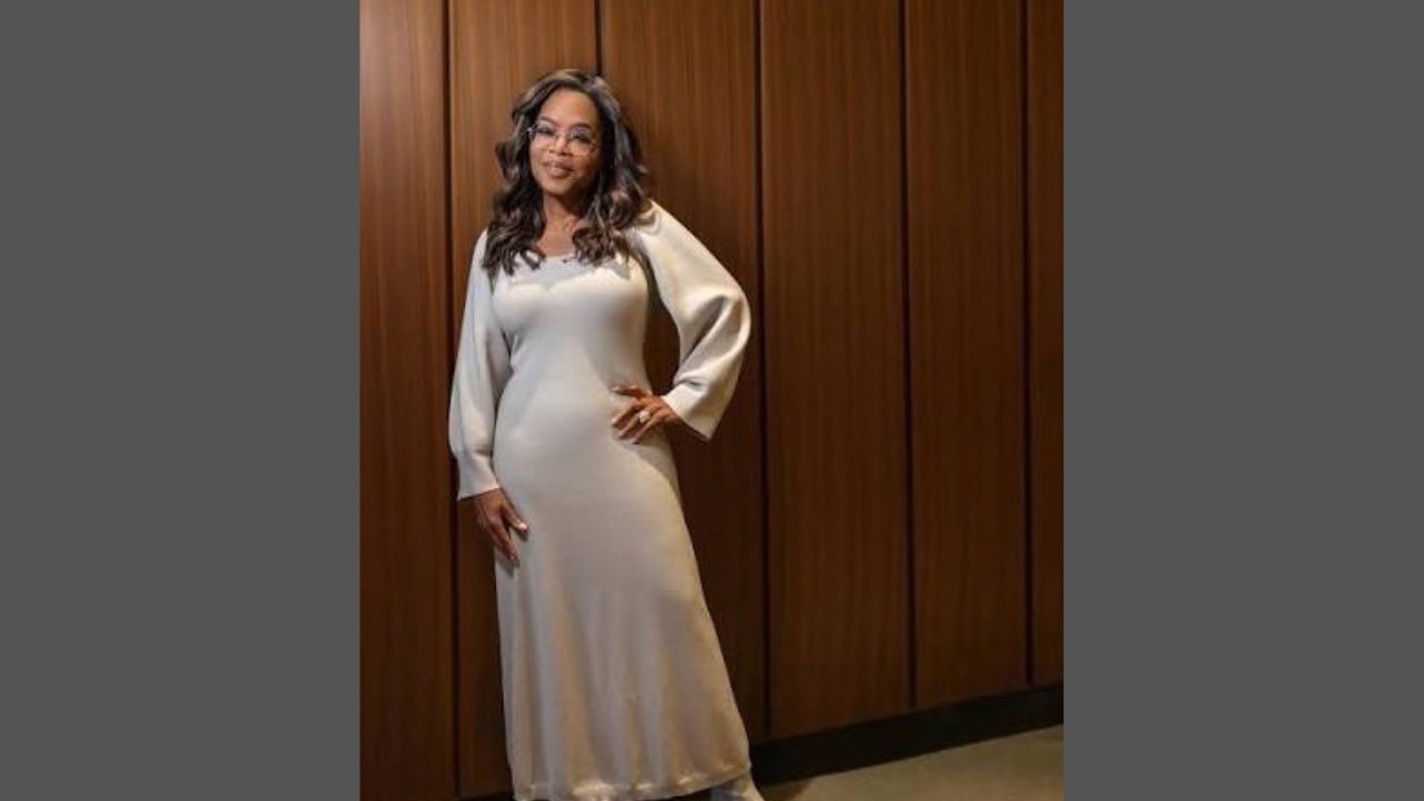 Oprah Winfrey in a long dress stands beside a rustic wooden wall, showcasing a serene and elegant pose.
